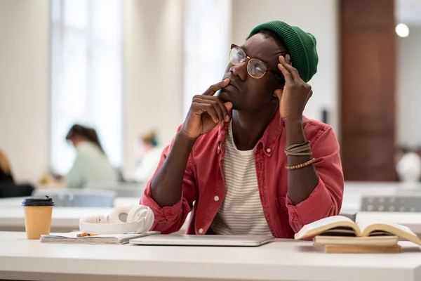 迷迷糊糊的非洲裔美国学生把目光投向一边 陷入沉思 在大学图书馆学习 坐在桌旁拿着笔记本电脑和课本 有选择性地集中注意力 教育和学习拖延 — 图库照片