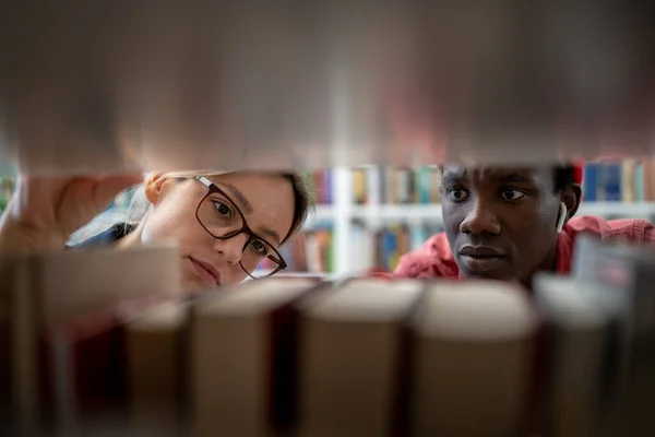 アフリカの男と白人の少女が図書館で一緒に勉強し 本棚の間に立って研究プロジェクトのためのソースを検索します 高等教育における共同学習 — ストック写真