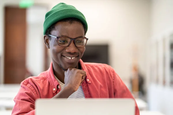 Cheerful happy African guy student looking at laptop screen while studying in college library, selective focus. Positive young black man enjoying e-learning, watching interesting webinar on computer