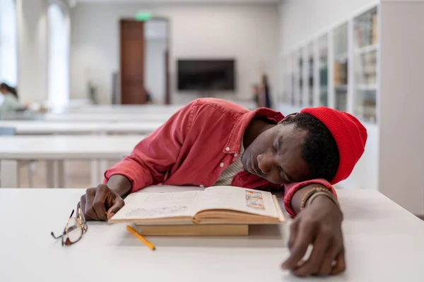 Exhausted African guy student fall asleep while studying in library, lying on desk with textbooks in classroom, taking nap during study, selective focus. Sleep, learning, and memory. Study burnout