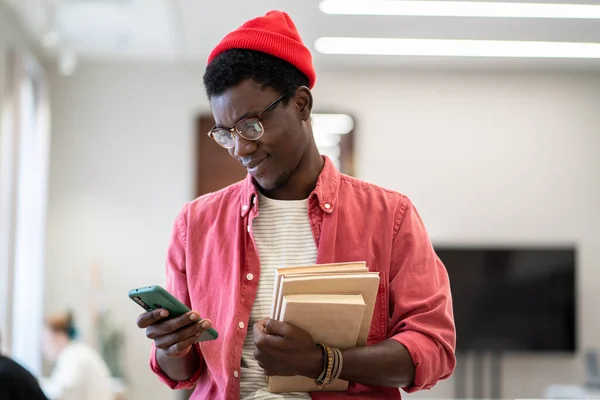 在图书馆学习时 微笑着拿着智能手机阅读短信的非洲裔美国学生 有选择地聚焦 在移动设备上使用调度应用程序的黑人男教师手持成堆的图书 — 图库照片