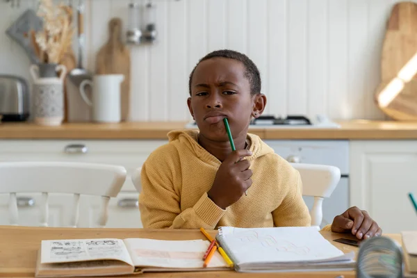 stock image Pondering focused child boy look at camera sit at home table with pencil near mouth thinking about difficult school tasks. Puzzled pensive kid schoolboy pout lips hold conceived some idea for lesson