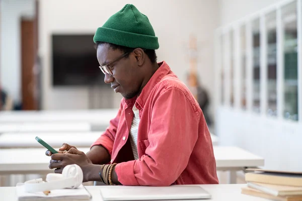 大学図書館のテーブルに座っているアフリカの学生の男は 勉強から休憩を取ってスマートフォンを持っています インターネットサーフィンや友人とチャット 勉強中の携帯電話の気晴らし — ストック写真