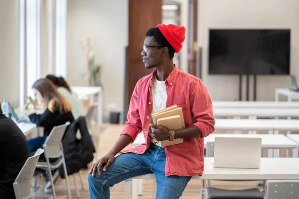 現代の大学図書館にノートパソコンを持って机の上に座り 教科書を持って窓の外を眺めながら勉強している黒人学生 学習環境と教育概念 — ストック写真