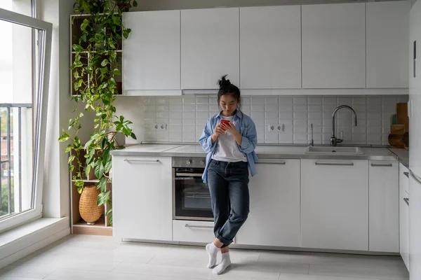 全长的亚洲妇女家庭主妇拿着手机 用应用程序支付公用事业费 千禧年女孩站在厨房里看着智能手机屏幕搜寻烹饪食谱 — 图库照片