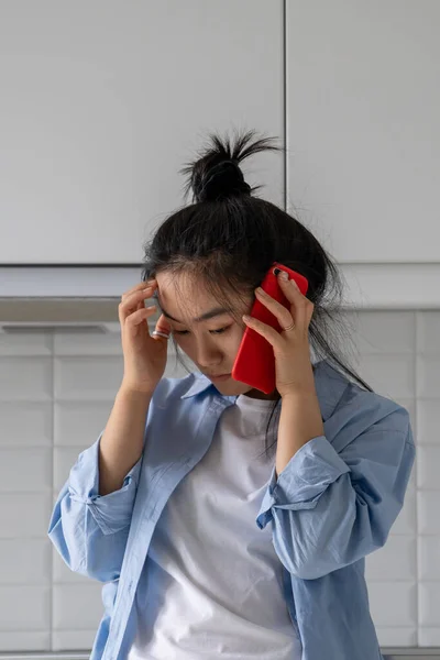 Sad disappointed Asian girl listening bad news over phone while standing in kitchen at home, feeling concerned, puzzled young woman holding cellphone having unhappy conversation