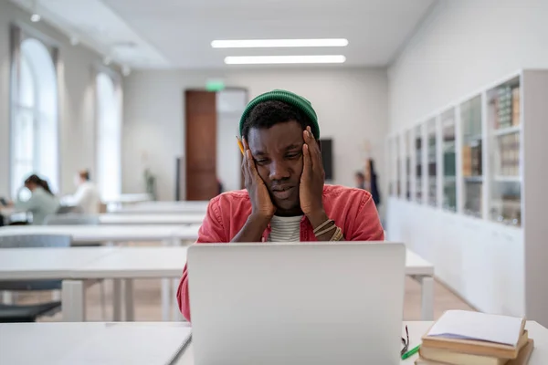 教育と精神衛生 ストレスの多いアフリカ系アメリカ人の男学生疲れています学習の座ってで大学図書館手に頭を保持し 欲求不満顔の式とラップトップを見て — ストック写真