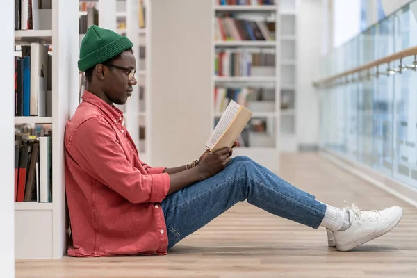 Focused Black student man book lover spending free leisure time in library, African American guy with book in hands sits on floor near bookcase enjoying reading. Hobby, education, university concept