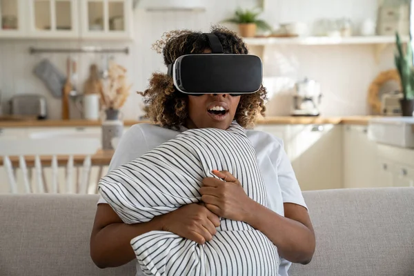 Afraid African American woman in virtual reality headset sits on home couch feeling excited, overwhelmed by watch horror movie. Affected black young female in 3D glasses hold pillow from feared game