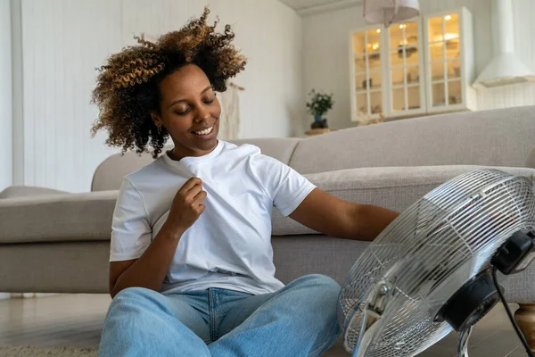 心满意足的快乐的非洲裔美国女人用电扇在凉爽的空气中让自己冷静下来 她讨厌闷热和闷热 快乐的微笑的女性坐在地板上 控制着来自家用通风设备的气流 — 图库照片