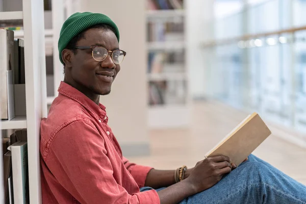 黑人学生男子图书馆访客手拿书坐在书架旁的地板上 看着相机 面带微笑 非裔美国人文学爱好者在公共图书馆的内部 阅读爱好 — 图库照片