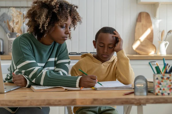 Focused biracial mother explaining difficult school task to attentive son for effective learning. African American personal tutor woman and little schoolboy sit at desk in home kitchen study together