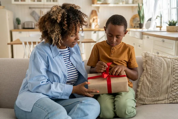 Caring loving African American mom greeting surprised small boy with present gift. Kind black woman give on birthday celebrate wrapped box with red ribbon to anticipating son sitting on couch at home
