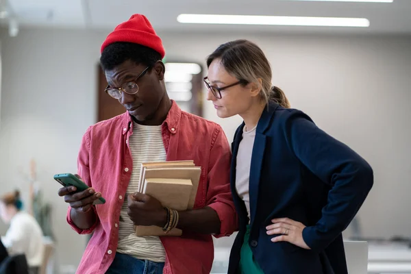 Two diverse students using mobile phone scrolling social media during study break, standing in classroom, study buddies Caucasian girl and African guy resting after studying together in library