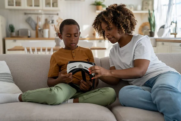 stock image African American mother showing son vr headset to son, while resting together at home, sitting on sofa in living room. Mom explaining virtual reality to child. Immersive technology and kids