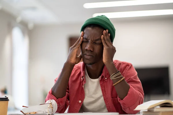 Stressed tired African American student man touching temples have headache after long time preparing for test in college library. Frustrated black hipster male feels nervous afraid of exam failure.
