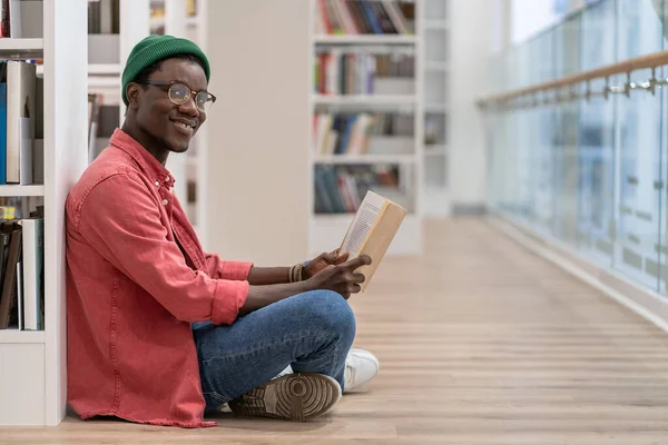 手に本を持って本棚の近くの床に座っている黒人学生図書館の訪問者 カメラを見て笑顔 公共図書館のインテリアでアフリカ系アメリカ人の男文学愛好家 趣味を読む — ストック写真