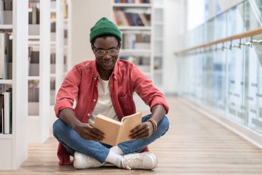 Kütüphanede yerde oturan siyahi öğrenci. Odaklanmış Afrikalı Amerikalı adam üniversitede okuyacak olan ders kitabı için malzeme hazırlıyor. Yüksek öğrenim, bibliyofil konsepti.