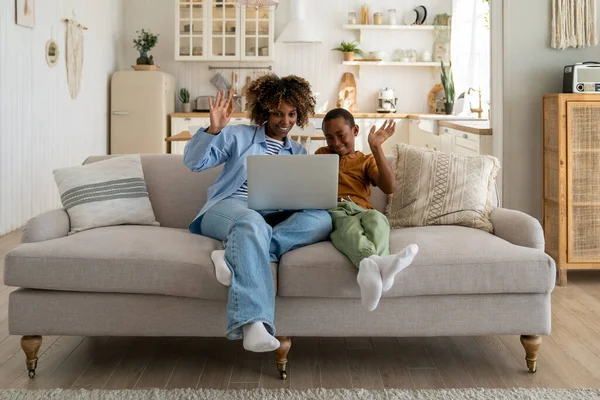 Happy family. African mother and kid son greeting online, waving hands, looking at web camera laptop for video call sitting on couch at home. Smiling mom and child having fun talking in video chat .