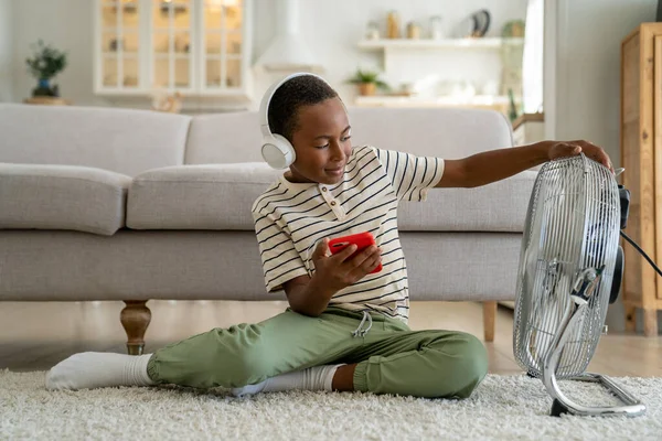 Interested smiling small african american child boy sit with smartphone in headphones on floor pleasure cooling with electric fan. Satisfied kid schoolboy use cellphone listen music chilling at home