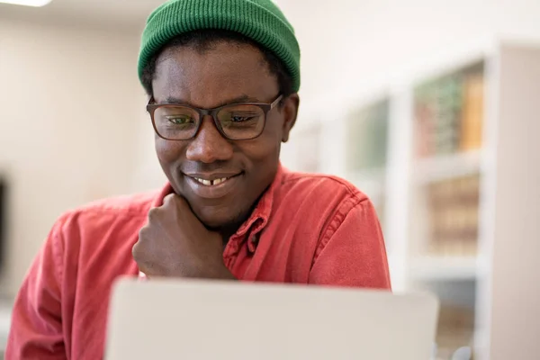 大学図書館で勉強しながら ラップトップ画面を見て陽気幸せなアフリカの男学生 選択的フォーカス 正の若い黒人男性楽しみます電子学習 コンピュータ上で興味深いウェビナーを見ます — ストック写真