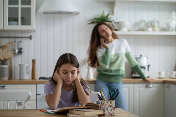 Concentrated teenage girl reading books preparing report or essay for school sits at table. Focused schoolgirl studying at home during quarantine sits in front of mother talking on phone