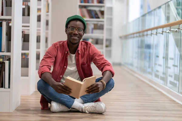Clever black student man with book sitting on floor in university library. African American guy smiling happily looking at camera, enjoy interesting effective studies in higher institution. Education