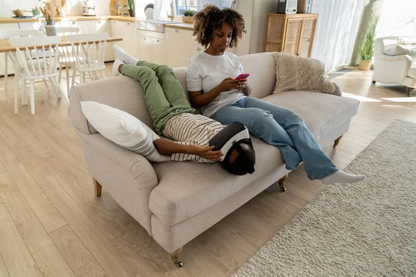 现代非洲裔美国人家庭的母亲和儿子们在家里的沙发上用数字设备休息 母亲一边坐在沙发上一边用手机一边和孩子们玩Vr游戏 家庭生活中的技术 — 图库照片