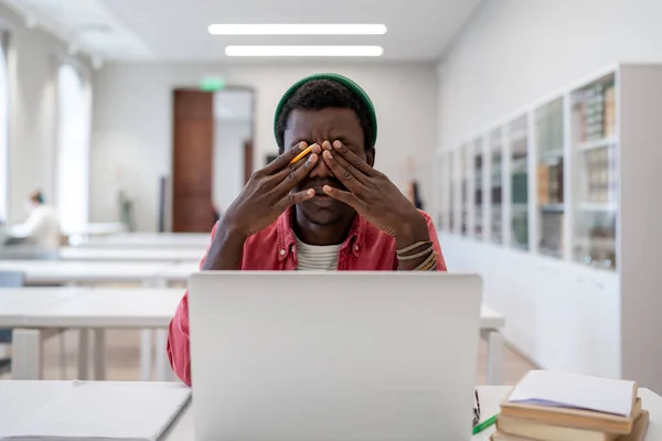 精疲力竭的非洲裔美国学生坐在图书馆的书桌前 手握笔记本电脑 感觉不舒服 擦干干巴巴的易怒的眼睛 试着集中精神 电脑工作后紧张偏头痛的疲倦黑人自由职业者 — 图库照片