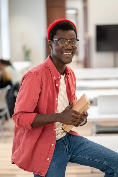 喜んでアフリカ系アメリカ人のヒップスター学生の男で眼鏡の図書館のテーブルの上に座っている本は親指を示すジェスチャー 推奨される効果的な大学教育のための手のような笑顔カメラを見て — ストック写真