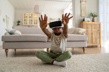 Yeni teknolojiler ve çocuklar. Sanal gerçeklik gözlüklü mutlu Afrikalı çocuk havaya dokunuyor, heyecanlı çocuk oturma odasında oturmuş VR oyunu oynuyor, evde metaevreleri keşfeden çocuk.
