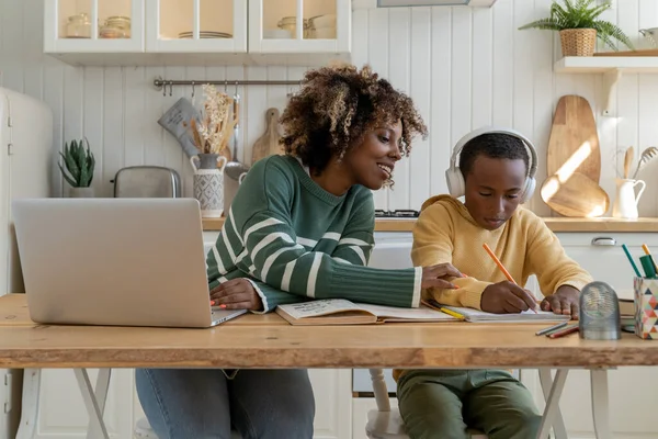 Pleased black mom freelancer works at laptop looks into son notebook focused on study in headphones. Home education test with tutor. Mother support child boy in learning of difficult school assignment