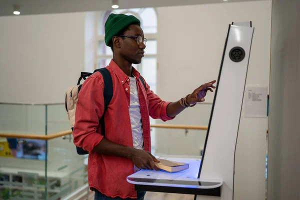Student African man using self-service electronic terminal to pay for goods in store without salesperson. Self-sufficient black guy standing indoors at college using touch screen vending machine