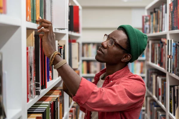 思慮深い集中アフリカ系アメリカ人のヒップスター男はビーニーの図書館に立つタッチ本棚大学の研究のための情報を見つける 焦点を当てた黒学生男で眼鏡検索で右の本 — ストック写真