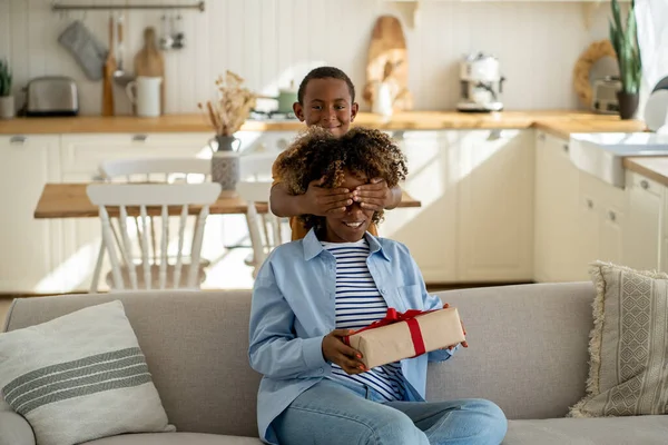 Happy Mothers Day. African American boy child making surprise for mother, covering eyes of surprised mom giving wrapped gift box, excited woman sitting on sofa receiving unexpected present from kid