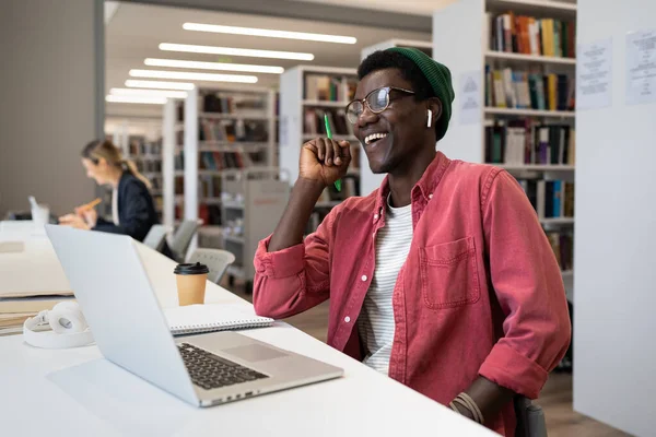 アフリカ系アメリカ人の学生がノートパソコンを持って図書館のテーブルに座り オンライン講義を見て笑っています 陽気な男はカジュアルなスタイルで身に着けているウェブセミナーやインターネットレッスンを見て楽しむ — ストック写真