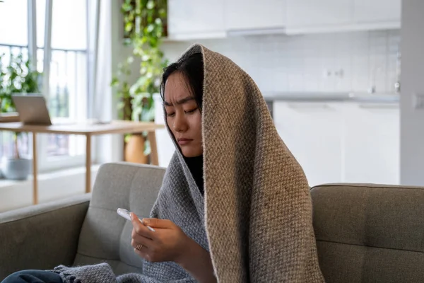 Upset unhealthy Asian Chinese young woman sitting under wool blanket on couch suffering from cold, looking at thermometer feeling sick unwell with high fever influenza virus illness cold flu migraine.