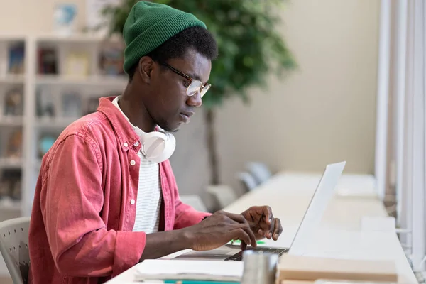 Young African American man freelancer using public library as workspace, typing on laptop. Focused black guy programmer or web developer wearing glasses working remotely on computer in coworking space