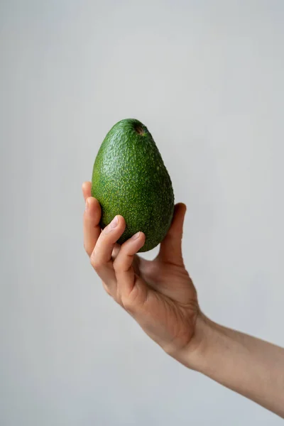 Human hand holding green ripe organic avocado against grey wall. Healthy vegetarian food. Nutrient-dense vegetables and fruits