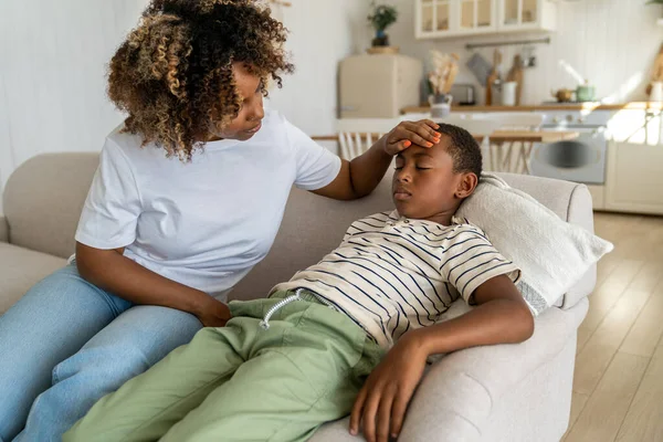 Worried caring African American mom touch forehead of sad child sitting on couch feeling unwell malaise nausea weakness. Attentive black woman mother check temperature of sick unhealthy schoolboy son