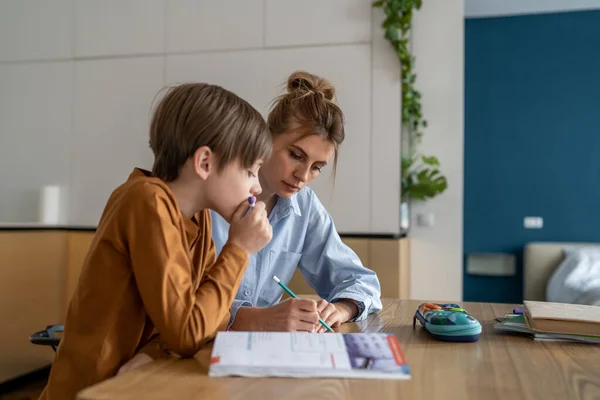 Young mother teaching teenage boy at home, mom helping puzzled kid son with difficult school task, supporting child in remote learning. Woman freelancer balancing parenting and remote work. Homeschool