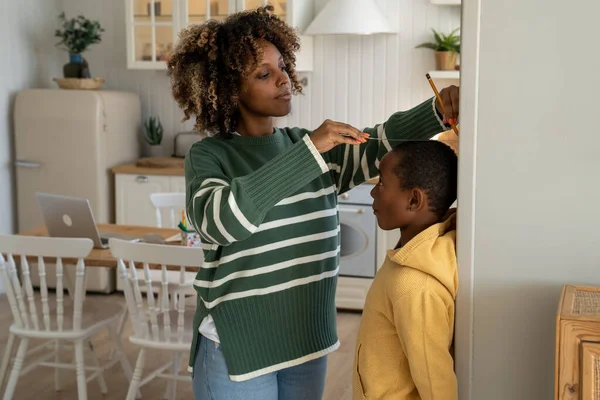 Caring african american mother measures changes height of child son on wall with pencil at home. Interested kid boy stand with mom wait for result hopes become taller tracking increase noting growth