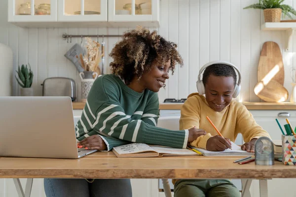 Joyful African American mom praises pleased son in headphones for success in studies and creative art task, distracted from remote work as freelance with laptop sitting together at home kitchen table