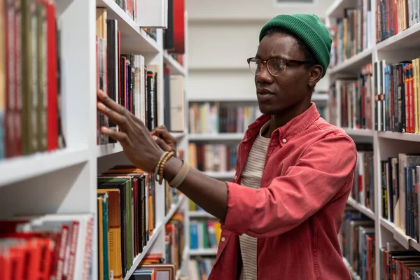 アフリカの男大学の学生は図書館の書棚の間に眼鏡をかけ 棚から本を拾い 研究プロジェクトを準備しながら文学を探します — ストック写真
