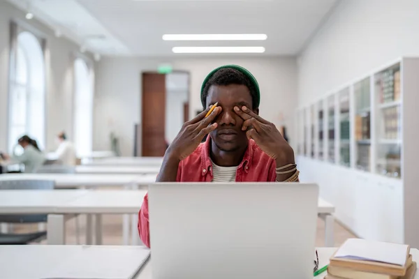 眼の緊張と仮想学習 疲れてアフリカ系アメリカ人の男大学の学生は ラップトップ感で机の上に座って圧倒され 図書館でオンライン学習中に画面から休憩を取ります — ストック写真