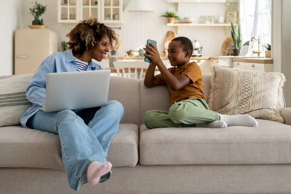 Happy African American family mother and son enjoy weekend at home rest on sofa with gadgets. Little boy child showing something on smartphone to busy mom freelancer, distracting her from remote job