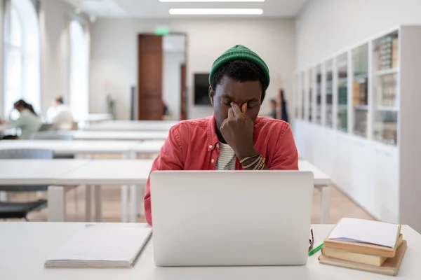 Academic stress. Tired frustrated black guy sitting in front of laptop in university library, suffering from digital eye strain caused by online education. Computer vision syndrome in students
