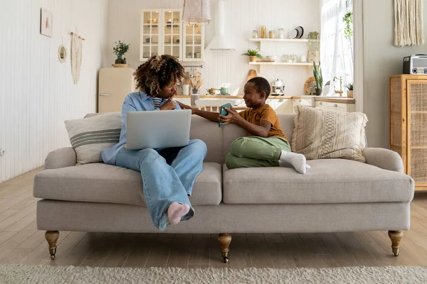 Motherhood and distance job. Busy African American mother freelancer sitting on sofa with laptop asking little son to be quiet during conference call, trying to manage remote work and child care