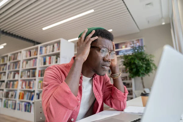 ストレスを受けたアフリカ系アメリカ人の学生は ラップトップの画面を見て眼鏡をかけて悪いテスト結果を受けています 図書館に座っている不幸な黒人は圧倒され あまりにも多くの家の仕事を持っている — ストック写真