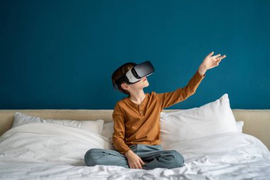 Sanal gerçeklik gözlüğü deneyimi yaşayan çocuk, fütüristik dünyaya dalmış. Akıllı telefon için VR gözlükleriyle video oyununda el havasına dokunan ilgini çeken küçük okul çocuğu yatağa otur.